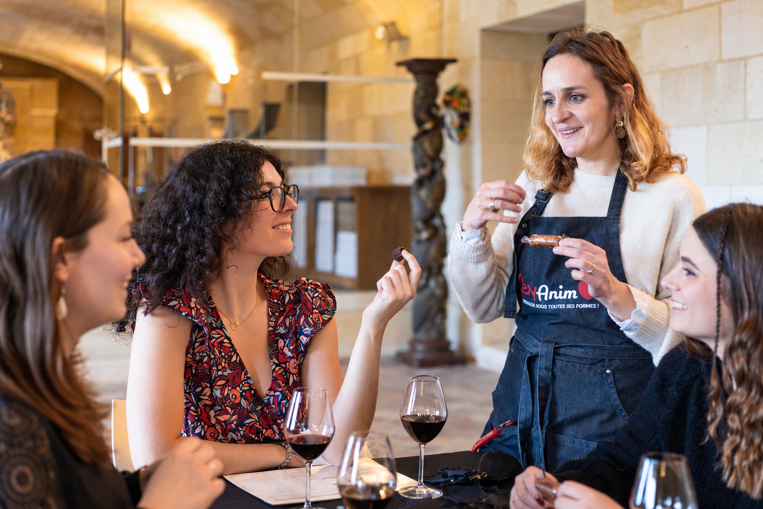 Une animatrice en tablier bleu tient un chocolat devant trois femmes souriantes attablées avec des verres de vin rouge, dans une salle voûtée en pierre.