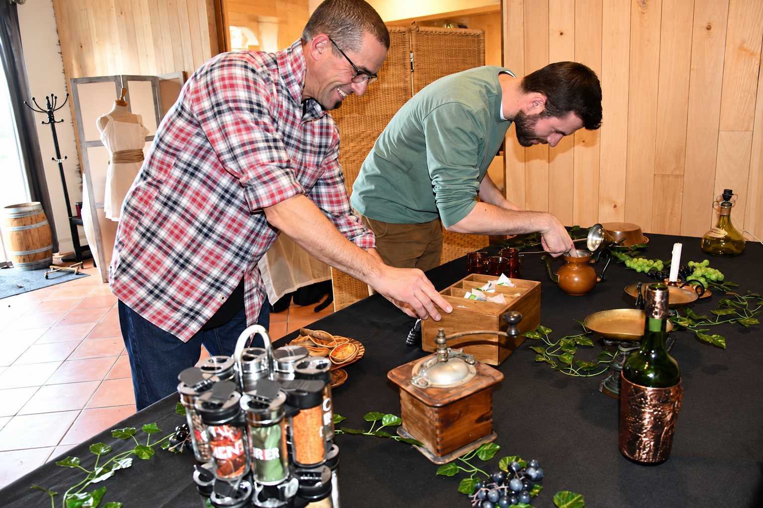 Deux hommes souriants disposent des objets anciens, un moulin à épices, des pots d’épices et des raisins sur une table noire ornée de feuilles de vigne.